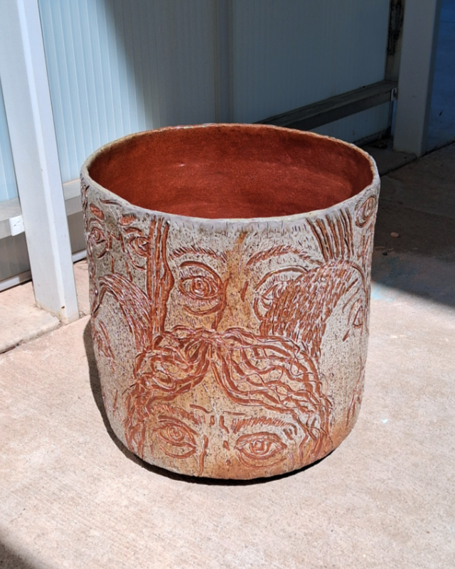 A ceramic planter by Dylan Cale Jones with faces incised its surfaces sits in the sun.
