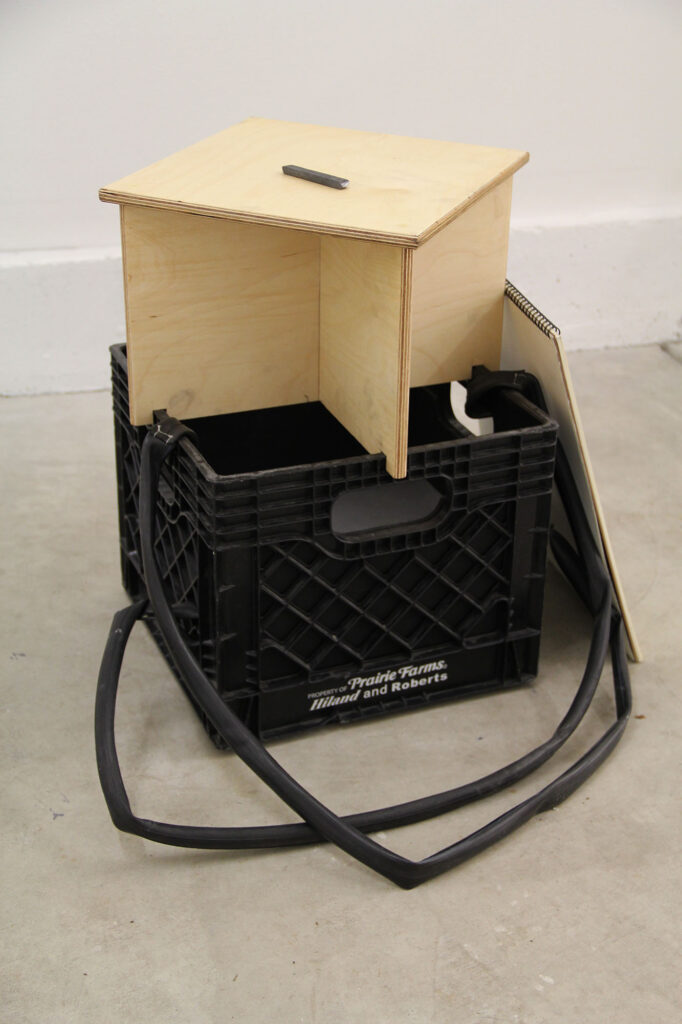 A plastic milk crate altered with shoulder straps for carrying and a wooden platform for sitting or standing