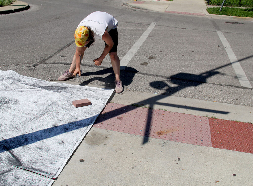 Dylan Cale Jones making a large-scale rubbing of the sidewalk underneath a police surveillance camera