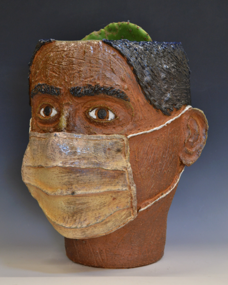 A ceramic sculpture of a head by Dylan Cale Jones . The head is facing slightly to the left. It is highly textured with wrinkles and is wearing a fabric medical mask. The top of the head sculpture is a planter, potted with a paddle cactus.